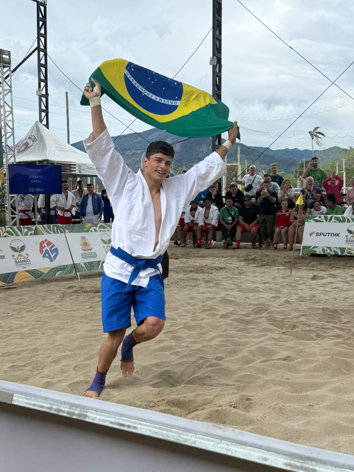 Atleta brasileiro comemora na areia erguendo a bandeira do Brasil durante o Mundial de Sambo de Praia 2026