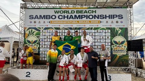 Equipe brasileira reunida no pódio do Mundial de Sambo de Praia 2026, em Caraguatatuba, ao lado da bandeira do Brasil e do telão oficial do campeonato