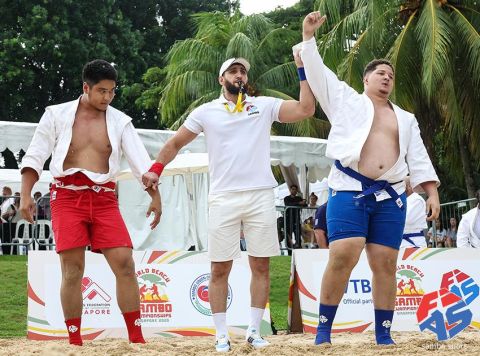 Árbitro levanta o braço do atleta vencedor de quimono azul após luta na areia do World Beach Sambo 2025