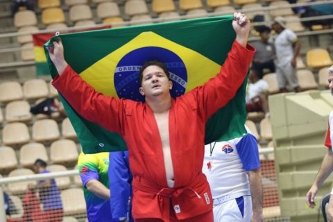 Atleta Vinicius Abreu, de quimono vermelho, comemora erguendo a bandeira do Brasil na arena