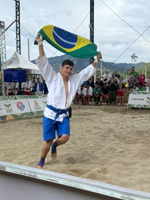 Atleta brasileiro comemora na areia erguendo a bandeira do Brasil durante o Mundial de Sambo de Praia 2026