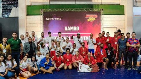 Grupo numeroso de atletas e técnicos com medalhas diante do painel de SAMBO do Arnold Classic