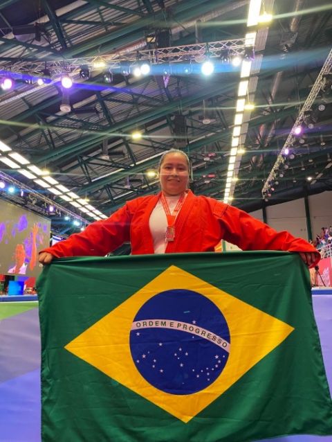 Atleta Priscila Rodrigues, de quimono vermelho, segura a bandeira do Brasil na arena de competição